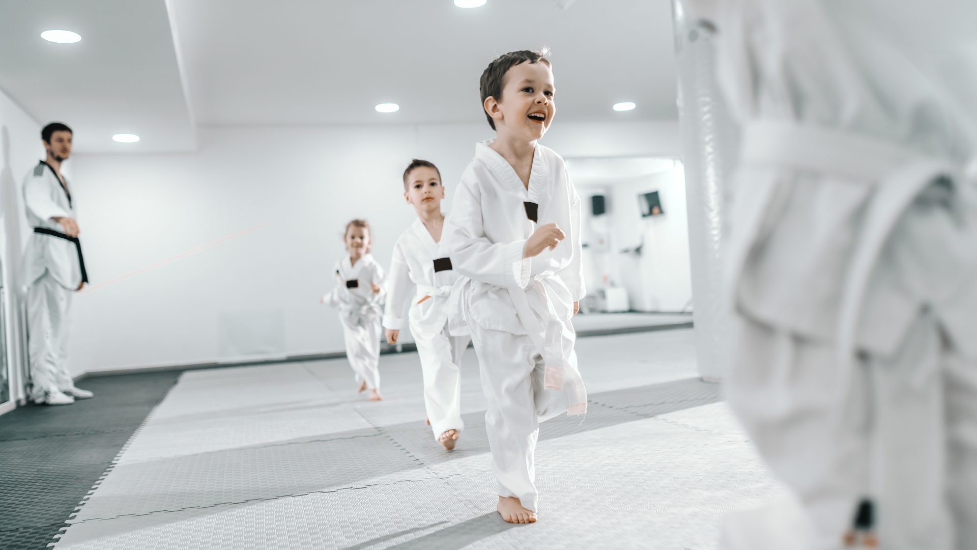 Des jeunes enfants s'exercent aux arts martiaux en courant sur un tatami devant leur entraineur.