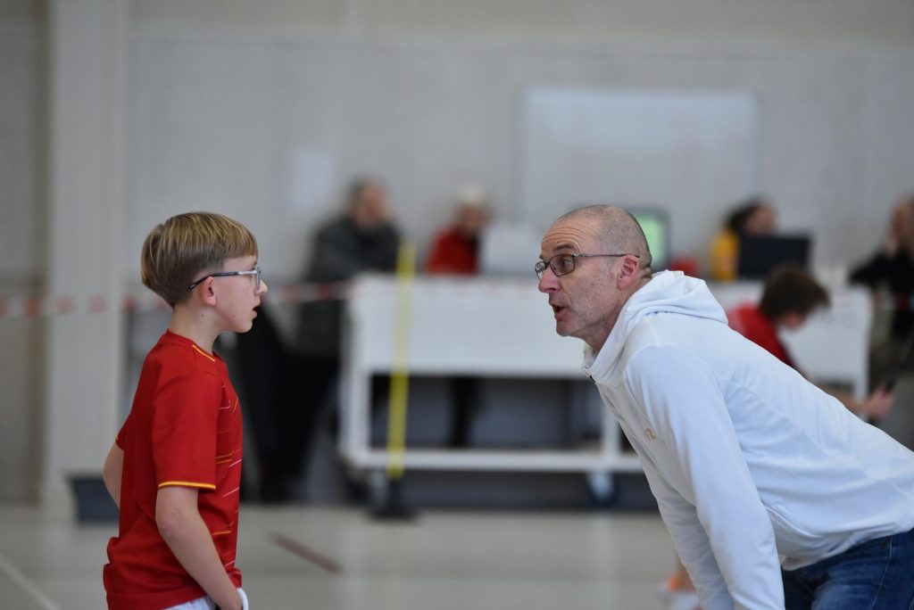 Un entraineur s'accroupit au niveau d'un jeune garçon pour lui donner des conseils de Badminton.